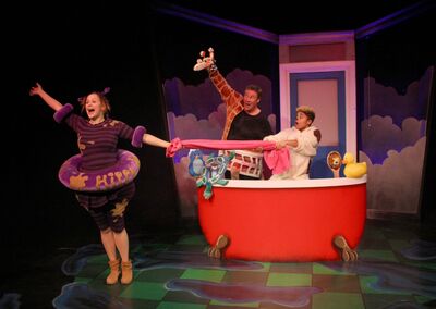 On stage there is a big red bath with two people standing in. One of them is pulling a pink sheet with someone standing out of the bath. The person out the bath is wearing a rubber ring and arm bands. She has her arms in the air and an excited expression on her face. There is a rubber duck and giraffe also on stage