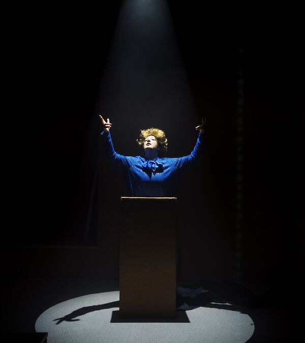 A performer on stage dressed as Margaret Thatcher stands at a podium with their arms outstretched