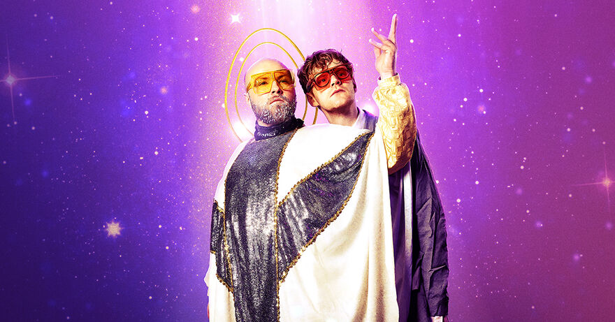 Two people standing, one wearing orange sunglasses and a white robe with glittery details. They are holding their arm up. The other person has dark orange sunglasses and is peering over the others shoulder. They are standing in front of a purple sparkly background