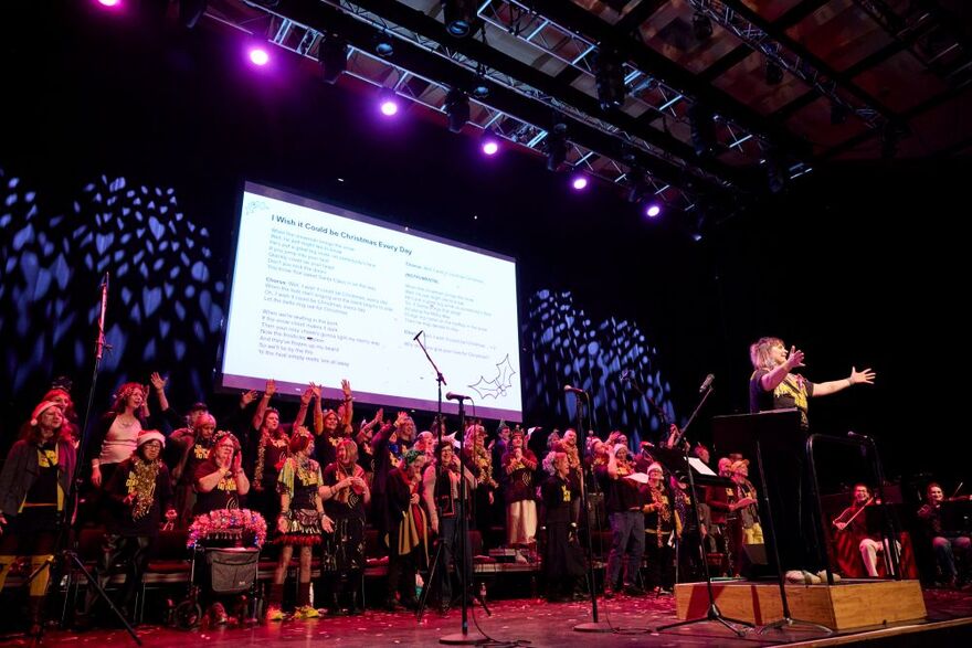 Loads of people celebrating on stage with confetti. They are wearing Christmads outfits including Reindeer antlers and tinsel. They are standing in front of a white background with Christmas song lyrics on it.