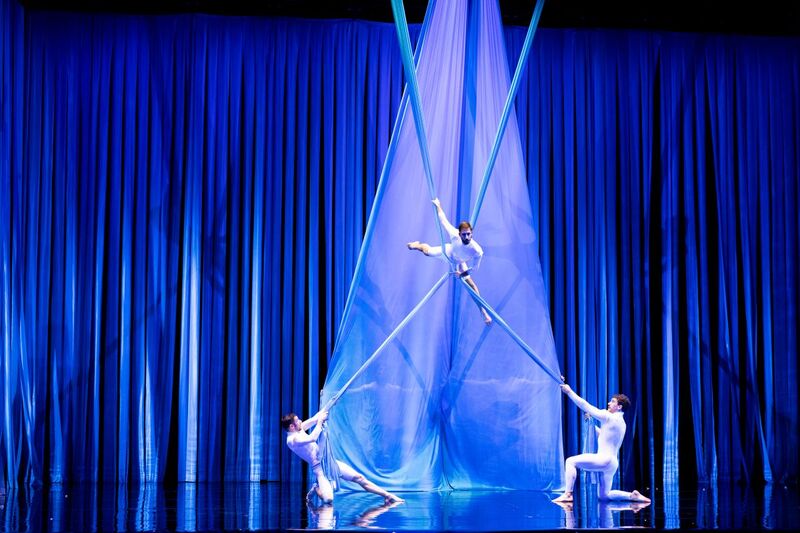 A theater stage is framed by deep blue curtains and illuminated with cool blue lighting. At center, a performer in a fitted white costume is suspended several feet above the stage on intersecting blue aerial silks, balancing with one leg extended outward and one hand gripping the fabric. Two other performers in matching white costumes kneel on opposite sides below, each pulling on the silks to create tension and support the aerial pose. The glossy black stage floor reflects the performers, fabric, and lighting, creating a striking, symmetrical composition.
