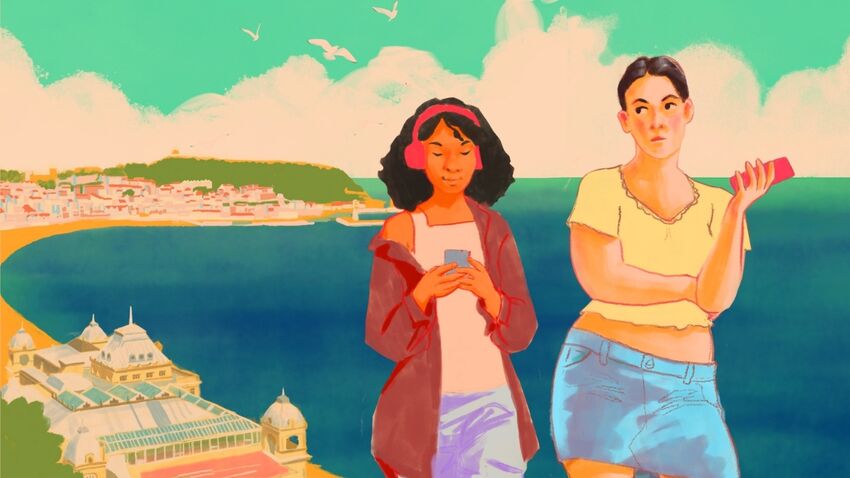 Two women standing next to each other with a view of the beach behind them. One is wearing headphones and looking at her phone. The other looks annoyed