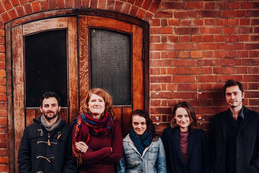 5 people standing in a line in front of a rustic brick wall. They're wearing warm, casual clothing and smiling at camera