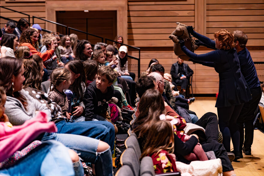 An audience of children and parents sit in stalls while two puppeteers stand at the front controlling a brown bear puppet