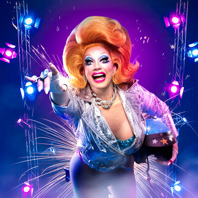 Ginger Johnson leaning towards camera with fireworks behind her as if she's 'blasting off'. She's wearing a big ginger wig, shiny silver jacket, white leather motorbike gloves and holding a motorbike helmet under one arm