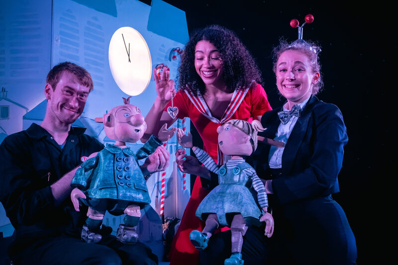 Two people hold wooder puppets of children with a girl holding a dangling heart between them