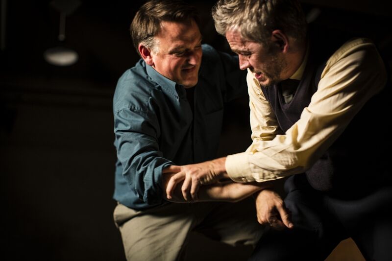Two men on a blacked out stage wrestle with each other hunched over