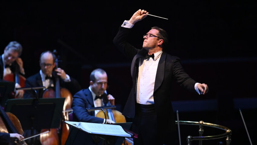A white, male conductor raises his arm, there's an orchestra visible in the background