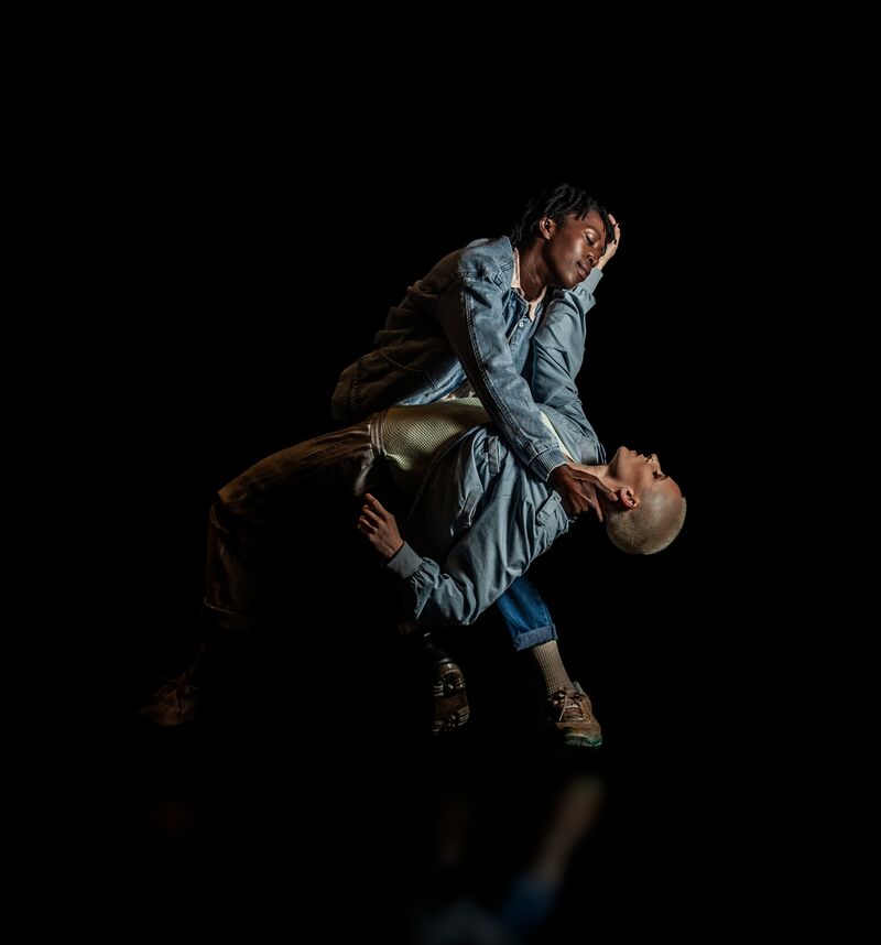 Two female dancers in blue denim jackets, One supporting the other in a deep backwards bend, both looking tenderly at each other. 