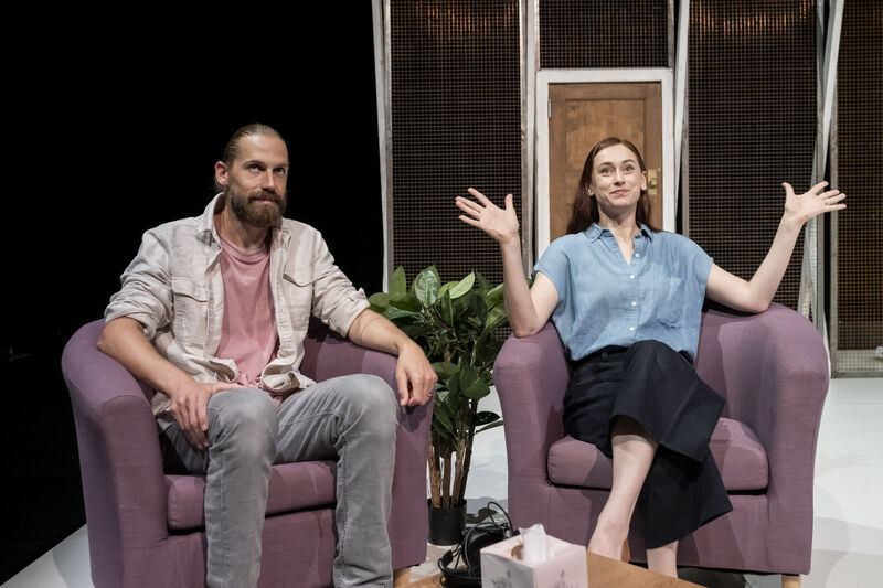A man and woman sit in purple armchairs. The woman has her arms raised in as if saying something sarcastically