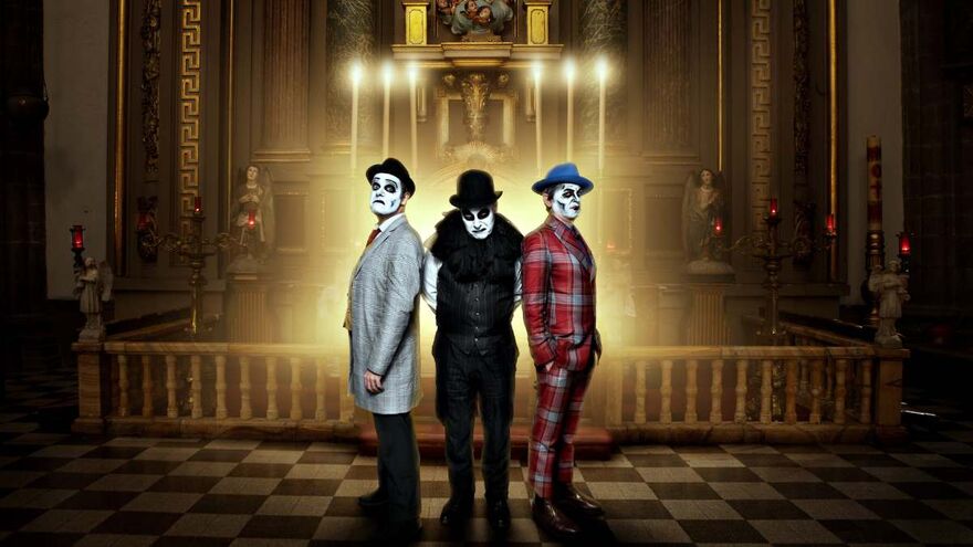 Three men stand side by side in front of a church alter with dramatic lighting. They are wearing suits (one grey, one black and one red tartan) and hats. All three have their face painted like skulls