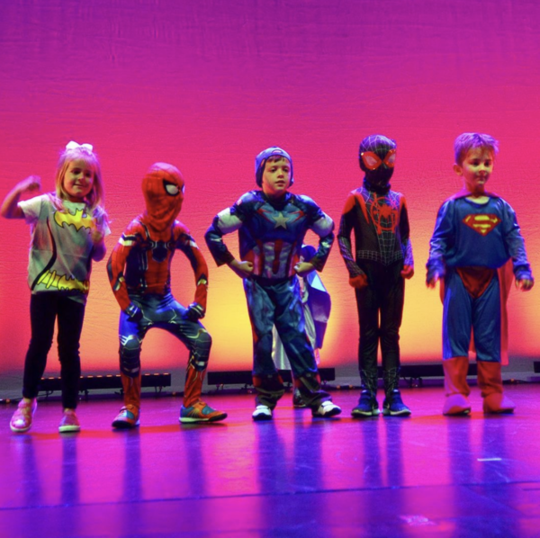 Children in super hero costumes pull silly poses in a line in front of a pink background