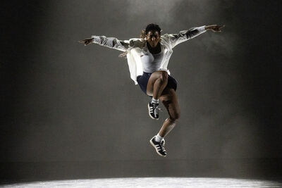 A female dancer in black and white clothes jumps with her arms spread like a bird under a single white spotlight