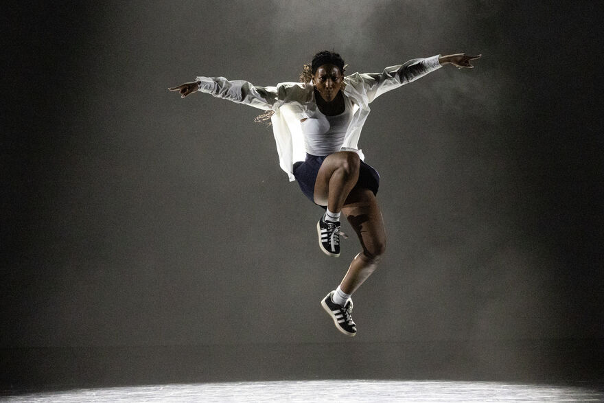 A female dancer in black and white clothes jumps with her arms spread like a bird under a single white spotlight