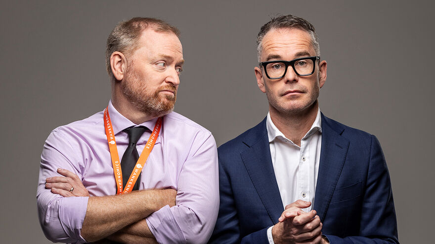Two men, with one with his arms crossed wearing a shirt and orange landyard looking at the other man. The other man has his hands together and staring at the camera