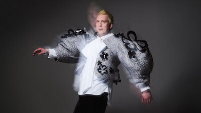 Joe Lycett stands against a dark background, with a big grey coat and a white shirt, looking directly at the camera