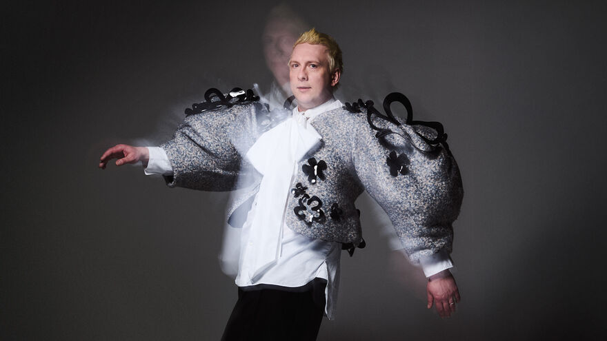 Joe Lycett stands against a dark background, with a big grey coat and a white shirt, looking directly at the camera