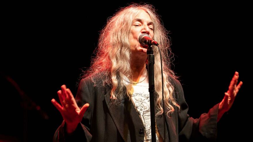 Patti Smith singing closely into a microphone on stage with her hands outstretched. She is a white woman in her 70s with long grey hair wearing a blazer over a graphic t-shirt