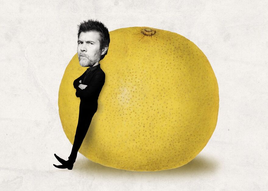 A small version of Rod Gilbert (a white man with short spikey hair, wearing a black suit and black shoes) stands leaning against a giant grapefruit, his arms folded.
