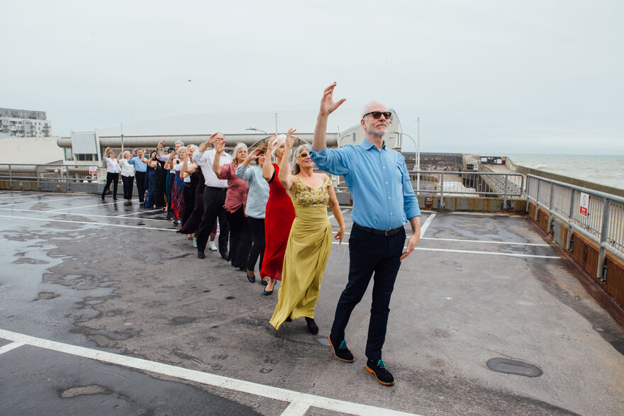 A group of older people stand in a line in a carpark, their arms are raised
