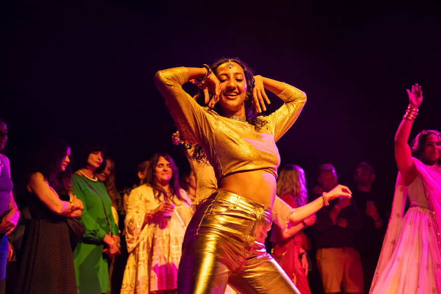 Women dressed in gold dancing in a room surrounded by happy people in different coloured clothing 