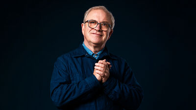 Harry Enfield standing with his hands together against a black background