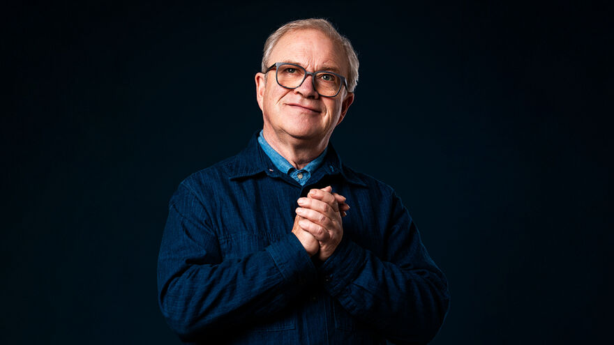 Harry Enfield standing with his hands together against a black background