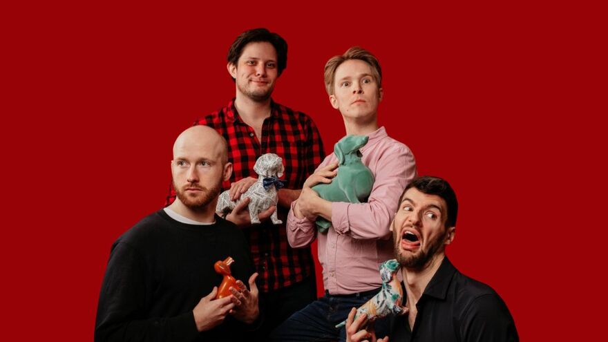 Four men holding paper mache dogs over a red background. Two of the men are kneeling with one looking to the side and another pulling a digusted face.  Two of the other men are standing behind, with one looking at the camera and the other looking ooncerned away from the camera