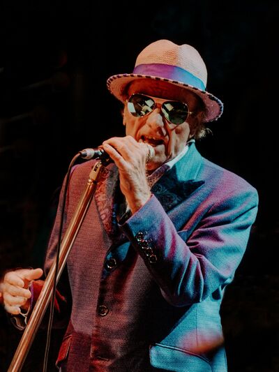 Van Morrison singing into a microphone. He is wearing a purple suit and brown hat and sunglasses