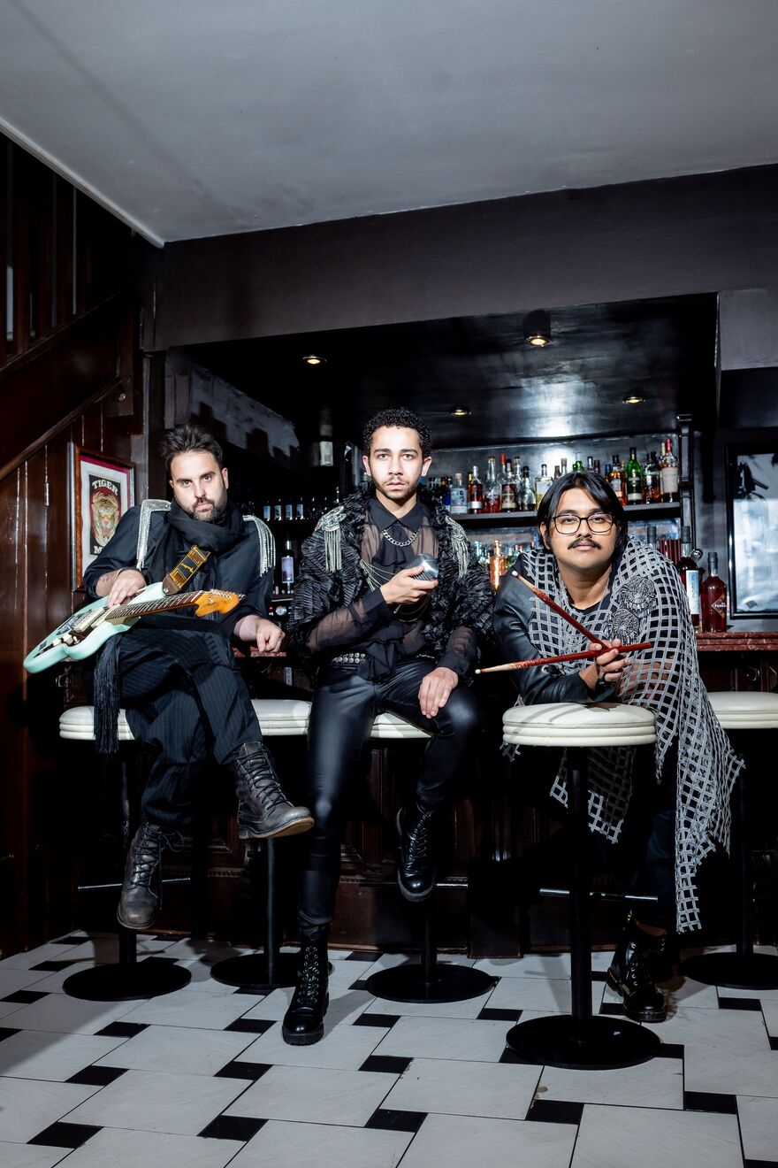 Three men sat on stools in a bar wearing layered black clothing. One is holding a guitar, one is holding a mic and one is holding drum sticks. Relaxed Performance icon