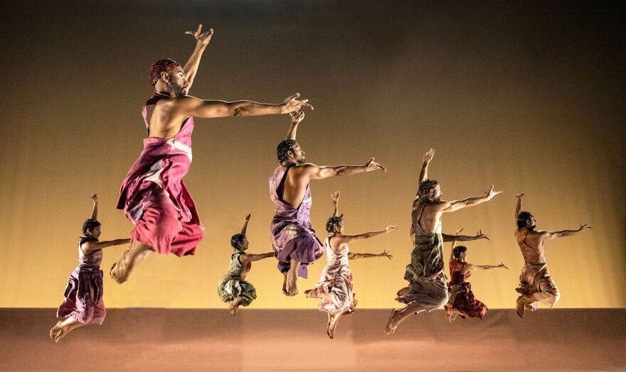 A group of dancers in flowing, colorful costumes leap across a stage in a synchronized contemporary dance performance, illuminated by warm golden lighting against a dark backdrop.