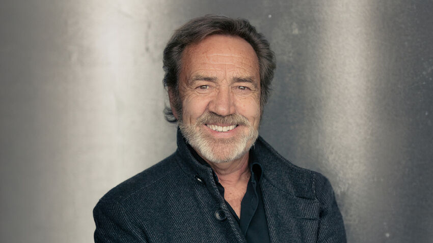 Robert Lindsay smiling at camera wearing a smart black shirt and grey coat