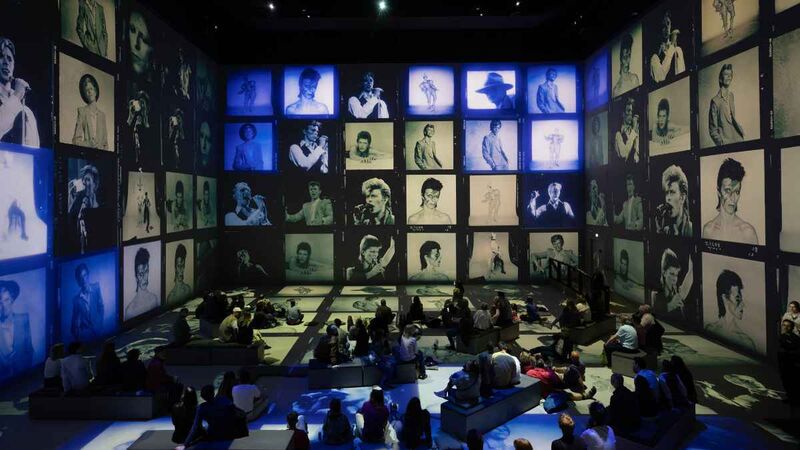 A dark immersive gallery space with dozens of projected black-and-white portraits covering the walls and floor, while visitors sit and stand in the center surrounded by blue and green lighting.