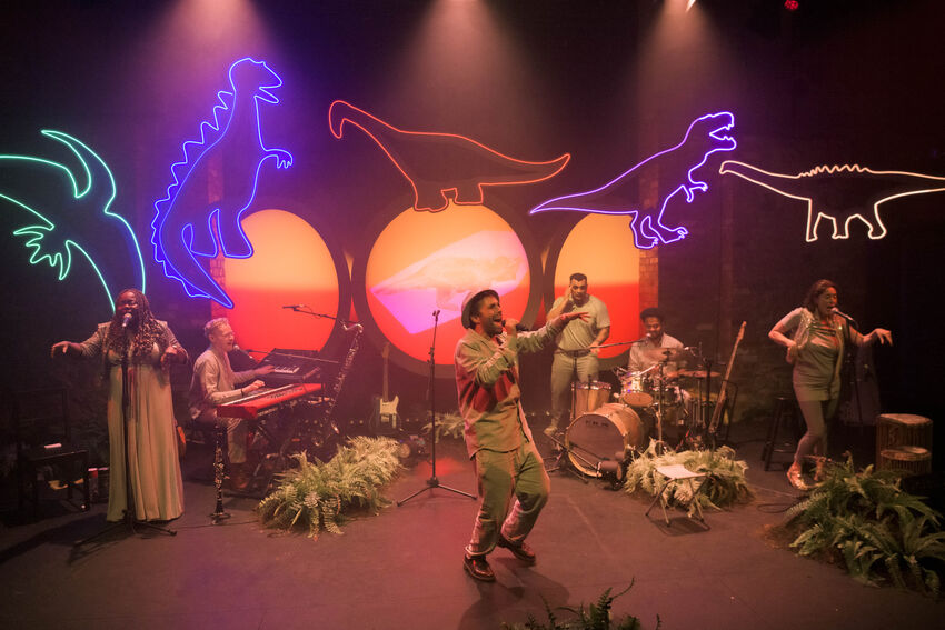 Five musicians playing and singing on stage under orange lighting with five neon dinosaurs lit up on the wall behind