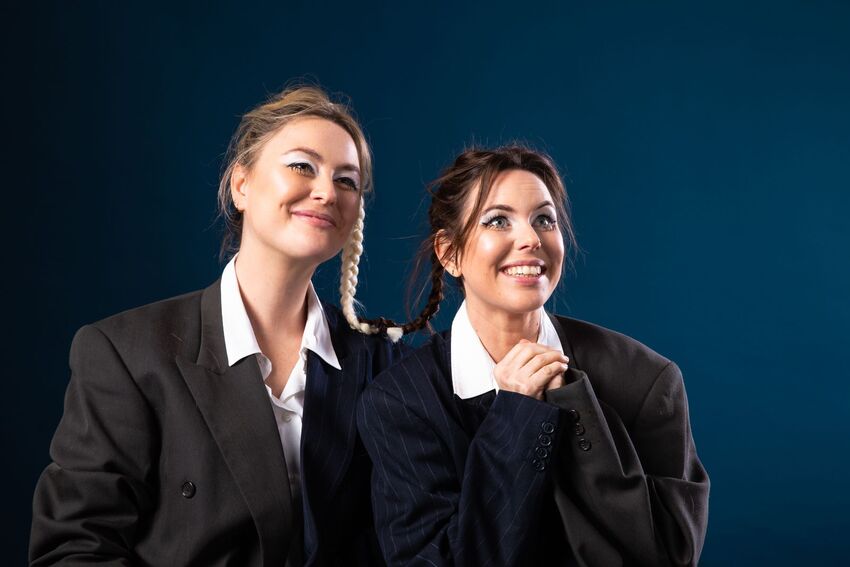 Two woman wearing black suits and white skirts are smiling. One of them is holding their hands together