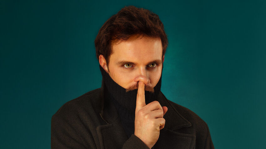 Vittorio Angelone over a blue background in a black coat, holding his finger up to his face 