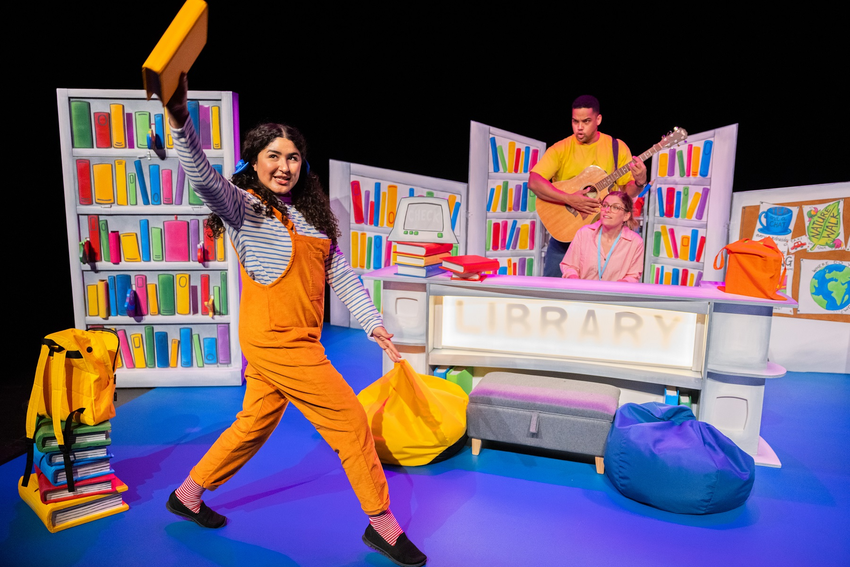 A colourful library set up on stage. A woman stands at the front wearing orange dungarees and carrying an orange book. Another woman sits behind a library desk wearing a lanyard. A man in the background is wearing a yellow t-shirt and playing an acoustic guitar