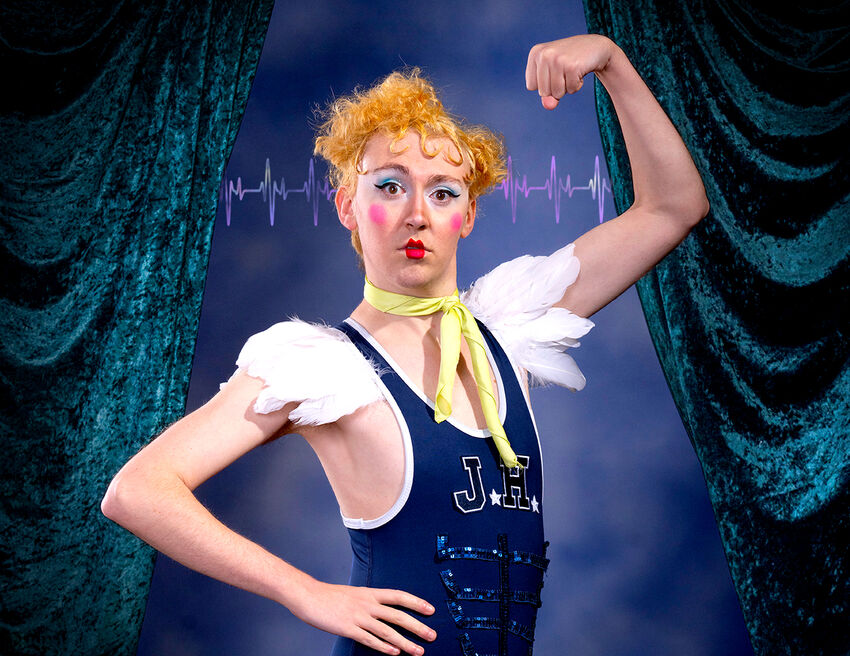 A person holding one arm up, with the other on their hip, wearing a blue vest and feather shoulder pads. They are wearing vibrant pink blush, red lipstick and blue eyeshadow 