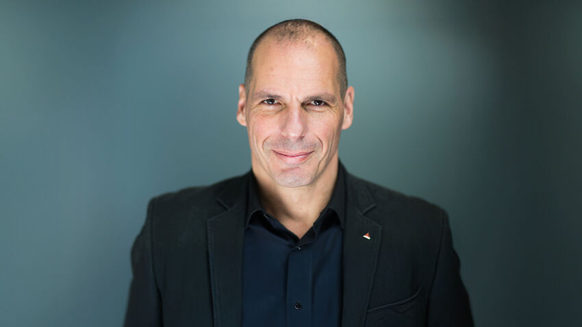 Yanis Varoufakis in a black shirt and blazer over a blue background