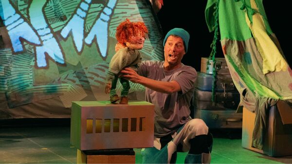 A performer interacts with a puppet on stage, surrounded by colorful sets and a projected backdrop.