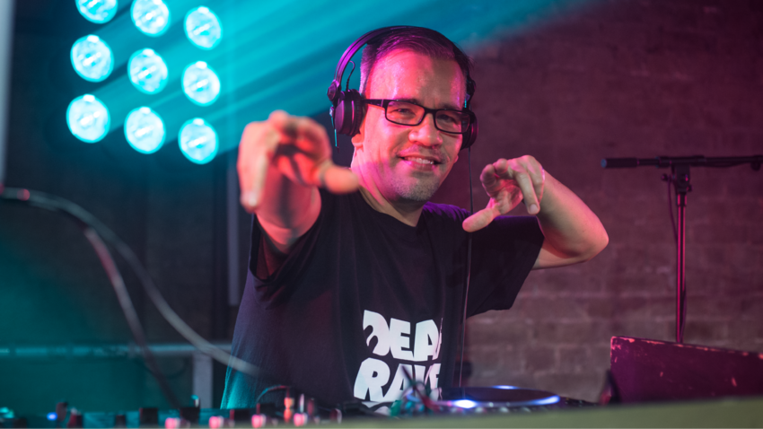 A DJ in a black t-shirt with headphones on. They are pointing at the camera