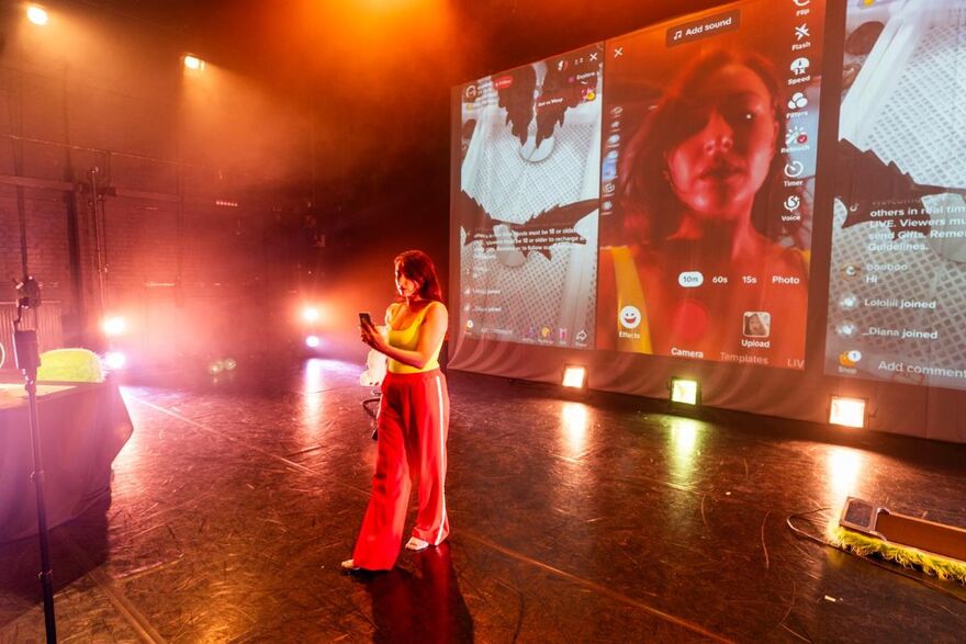 A woman on stage wearing red tracksuit bottoms and a yellow top. She is on her phone. Behind her is a projection of her phone on facetime, and other screens of tiktok. The stage is dimly lit with different spotlights