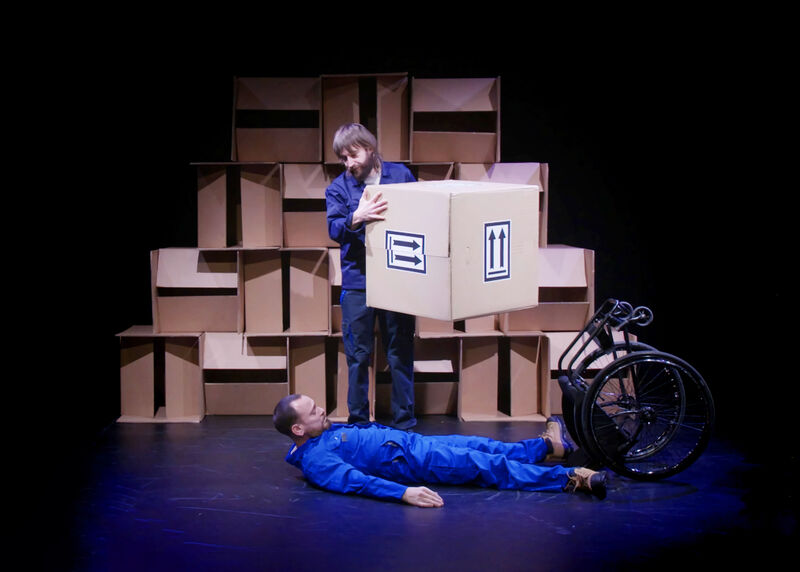 A man lay on the floor having fallen out of his wheelchair that is toppled next to him. Above him stands another man who is holding a big cardboard box with arrows on it.