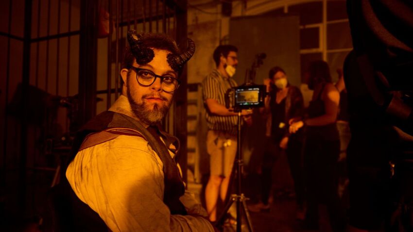 A man sits in the foreground with spooky makeup and devil horns. In the background there is a screen and a film crew