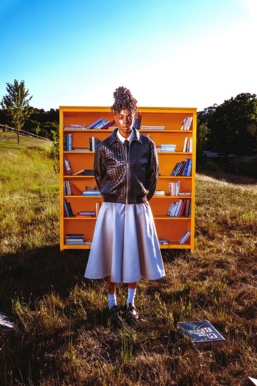 A woman wearing a blue dress with a brown leather jacket and a tie. She is standing in front of an orange bookcase in a field with trees
