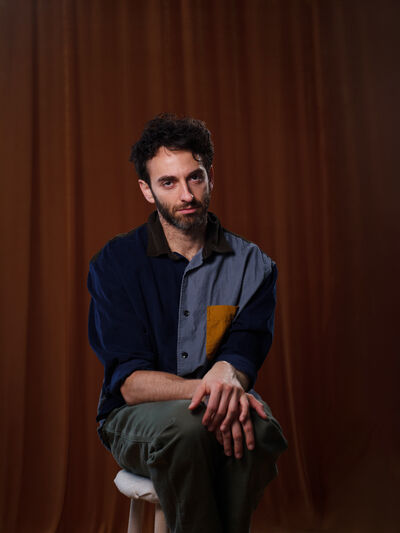 Gianmarco Soresi wearing a two-tone blue shirt with a mustard yellow pocket and dark grey trousers sitting on a stool with his hands on his knees in front of a dark brown curtain