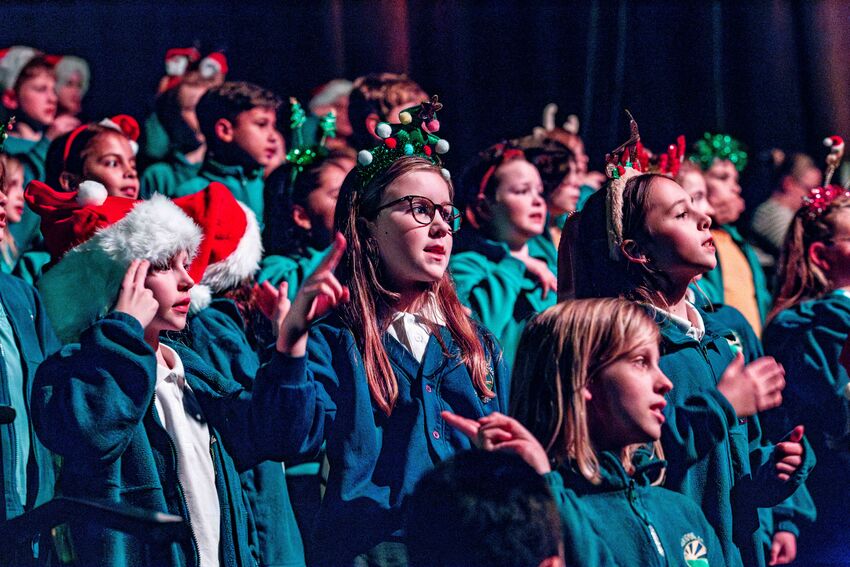 A group of school children are singing. They are wearing festive tinsel headbands and santa hats
