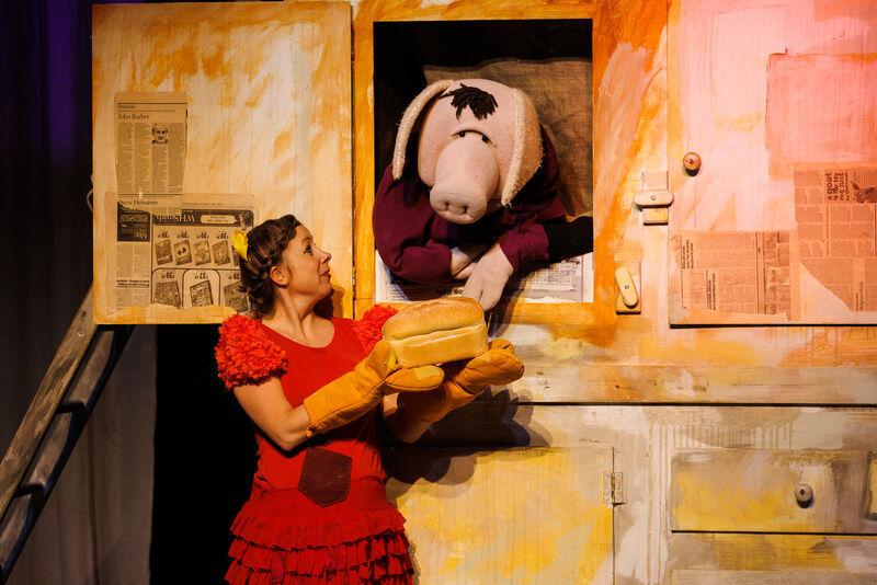 A large pig puppet leans out a cupboard towards a loaf of bread being held by a woman dressed as a red hen