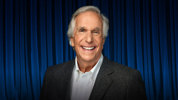 Henry Winkler is a white male in his late 70s smiling as he stands in front of a blue theatre curtain