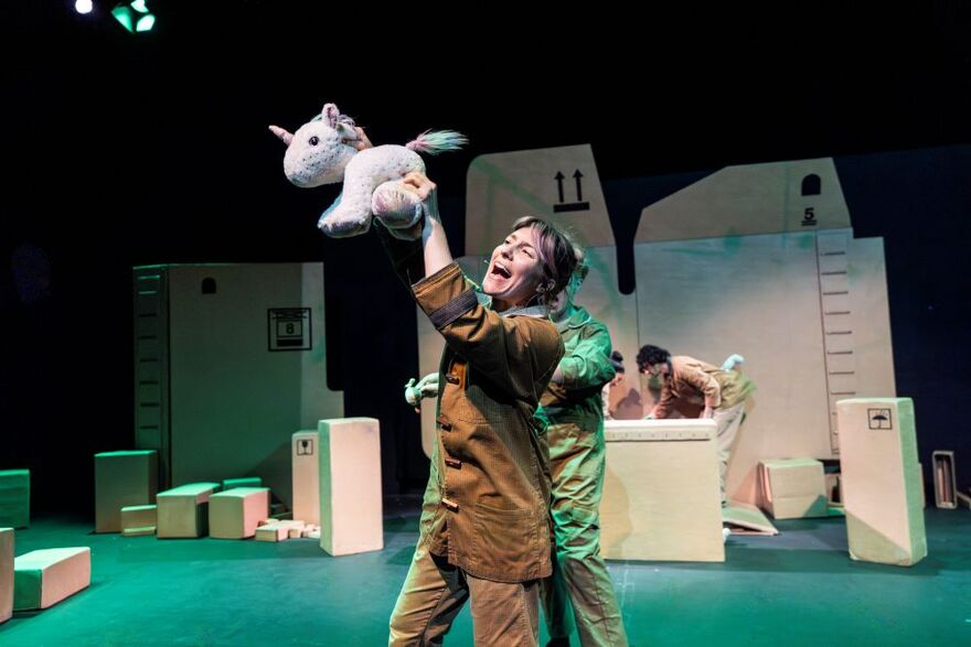 Someone standing on stage holding up a fluffy unicorn toy. She looks happy on stage surrounded by cardboard boxes.